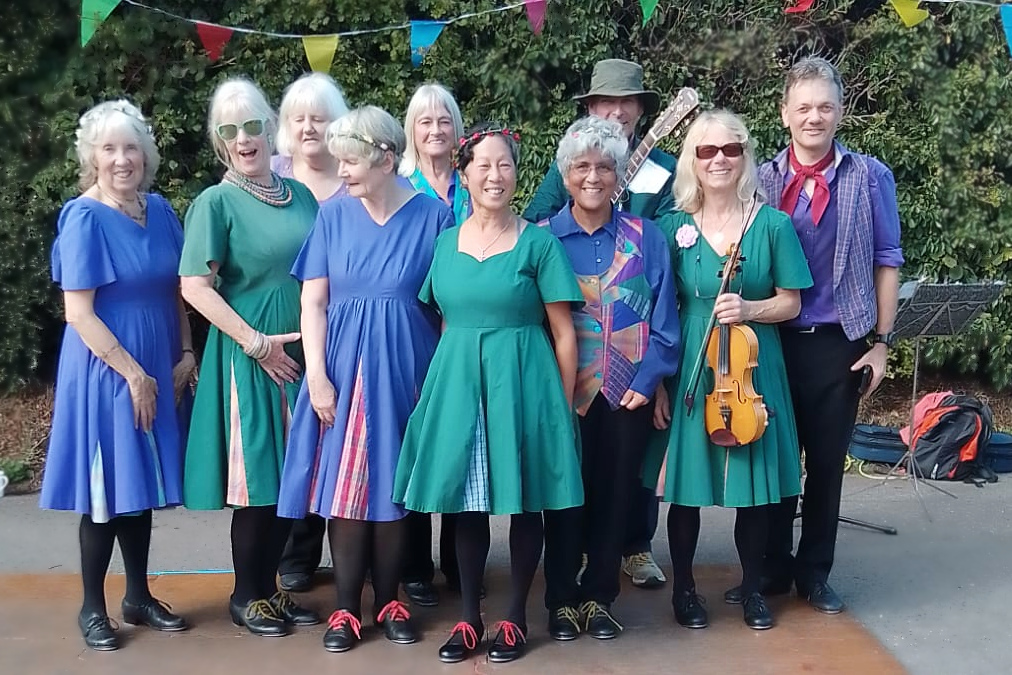 A group of dancers wearing green and blue outfits, one holding a fiddle.