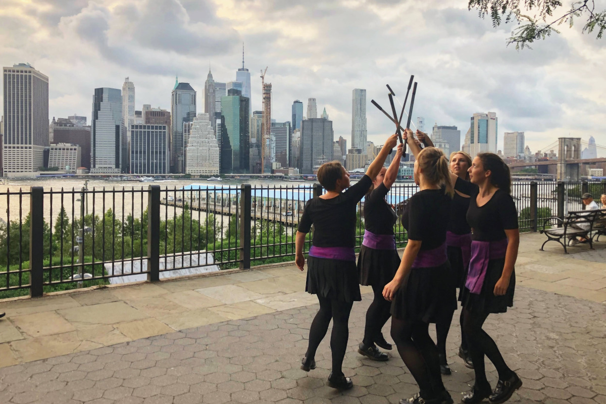 A group of dancers with sticks in front of a river and a city skyline.