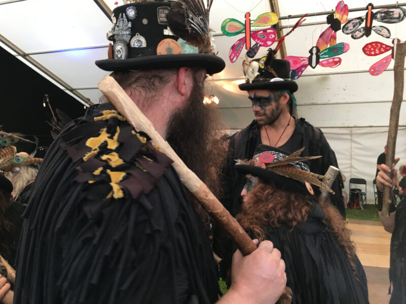 Three people wearing black costumes, one carrying a wooden stick.