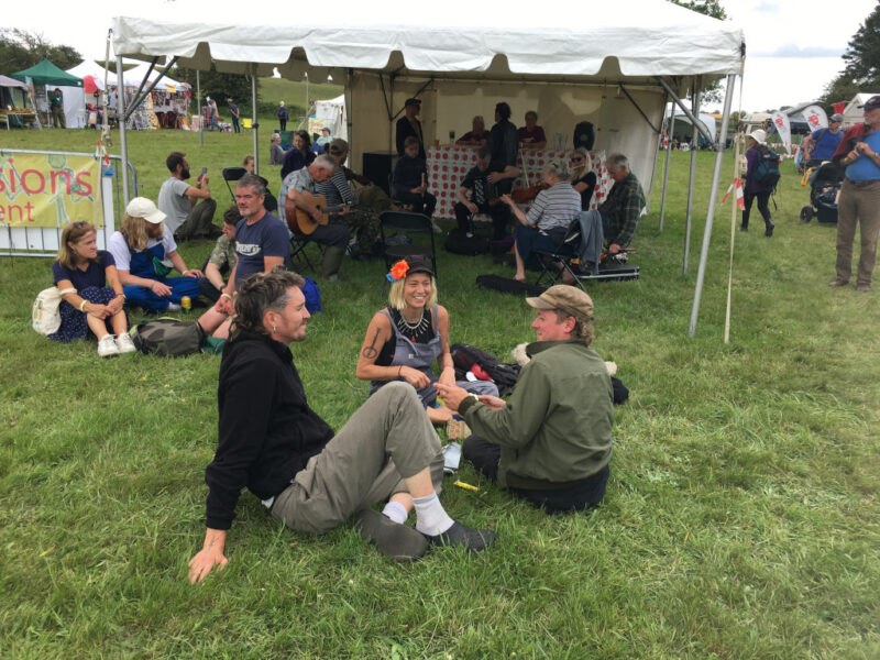 Three people sitting on grass with people playing instruments in a tent in the background.